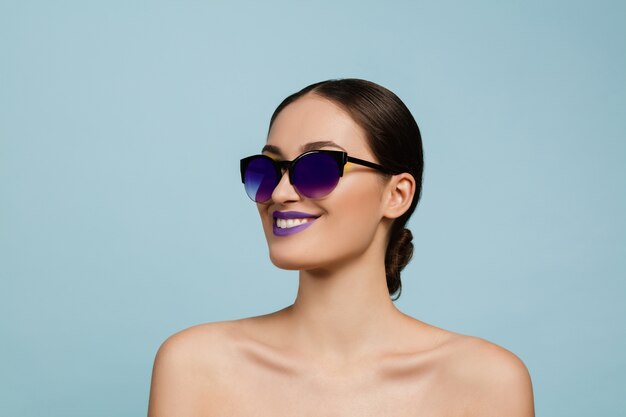 Retrato de mujer hermosa con maquillaje brillante y gafas de sol sobre fondo azul de estudio. Maquillaje y peinado con estilo y de moda. Colores de verano. Concepto de belleza, moda y publicidad. Sonriente.