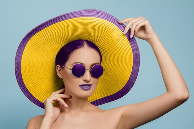 Retrato de mujer hermosa con maquillaje brillante, sombrero y gafas de sol en estudio azul
