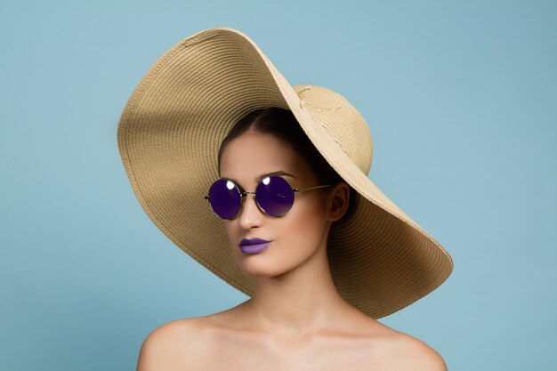 Retrato de mujer hermosa con maquillaje brillante, sombrero y gafas de sol en estudio azul