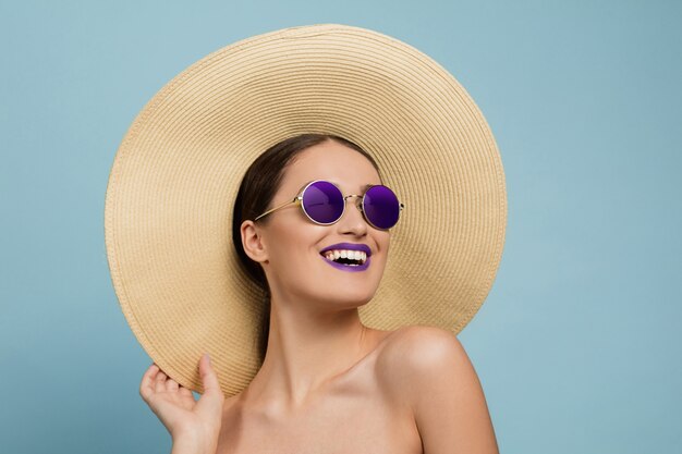 Retrato de mujer hermosa con maquillaje brillante, sombrero y gafas de sol en estudio azul