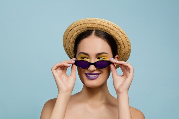 Retrato de mujer hermosa con maquillaje brillante, sombrero y gafas de sol en estudio azul