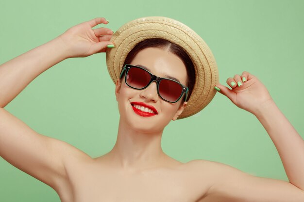 Retrato de mujer hermosa con maquillaje brillante, sombrero y gafas de sol en estudio verde