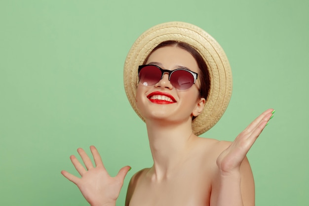 Retrato de mujer hermosa con maquillaje brillante, sombrero y gafas de sol en estudio verde