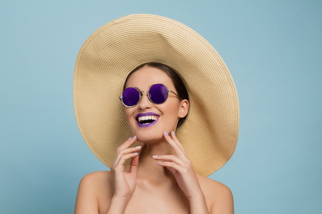 Retrato de mujer hermosa con maquillaje brillante, sombrero y gafas de sol. Hacer y peinado elegante y de moda. Colores de verano. Riendo.