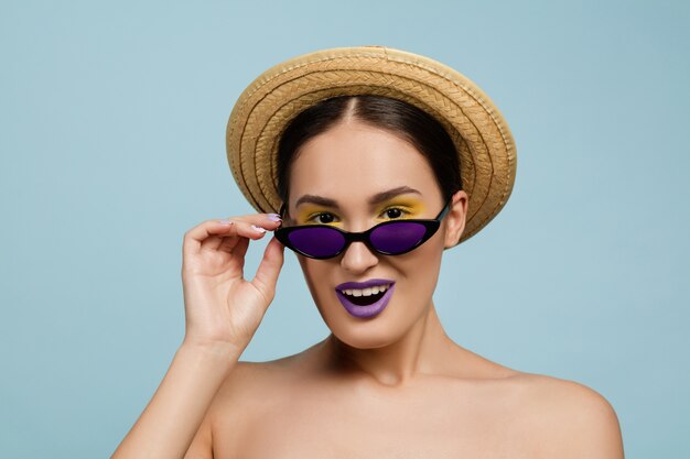 Retrato de mujer hermosa con maquillaje brillante, sombrero y gafas de sol sobre fondo azul de estudio. Hacer y peinado elegante y de moda. Colores de verano. Belleza, concepto publicitario. Busca gafas.