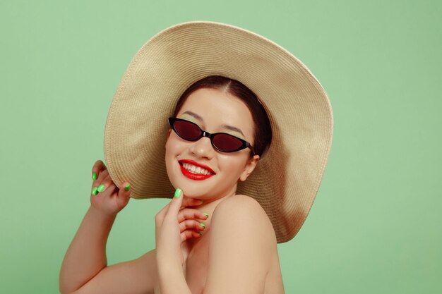 Retrato de mujer hermosa con maquillaje brillante, sombrero y gafas de sol sobre fondo verde de estudio. Hacer y peinado elegante y de moda. Colores de verano. Concepto de belleza, moda y publicidad. Sonriente.