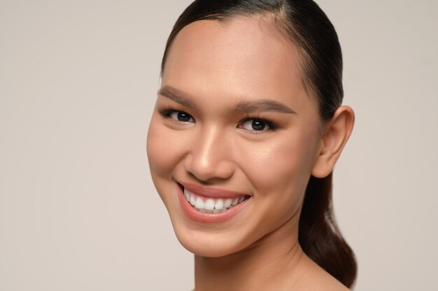 Retrato de mujer hermosa modelo con maquillaje diario fresco y sonrisa con dientes con rostro hermoso en pared gris en estudio