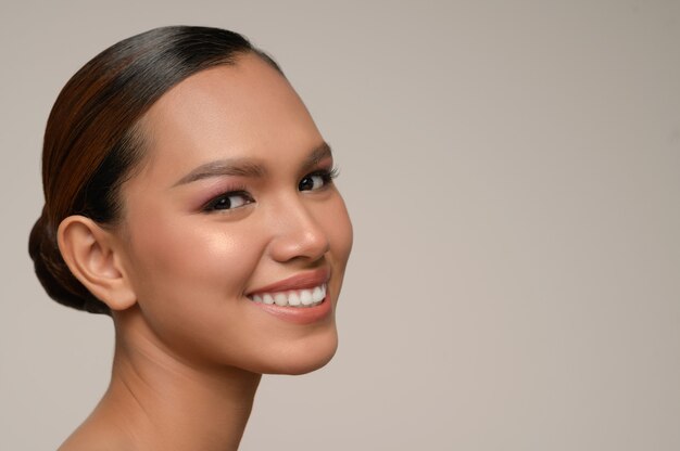 Retrato de mujer hermosa modelo con maquillaje diario fresco y sonrisas con dientes con rostro hermoso en pared gris en estudio