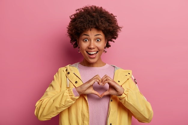 Foto gratuita retrato de mujer hermosa con peinado rizado hace gesto de corazón sobre el pecho, expresa amor, dice ser mi san valentín, tiene expresión positiva