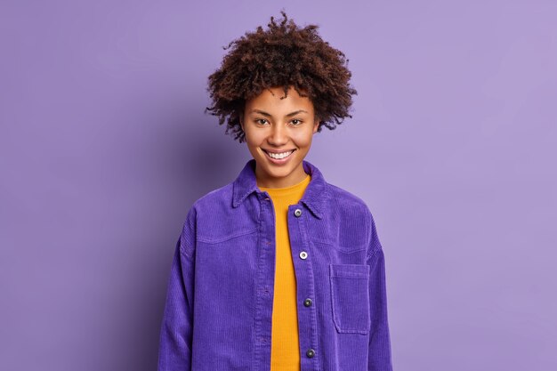 Retrato de mujer hermosa de pelo rizado sonríe alegremente viste una chaqueta púrpura de moda en un tono con el fondo se siente encantado posa alegre charlas interiores con un compañero de trabajo Concepto de emociones positivas