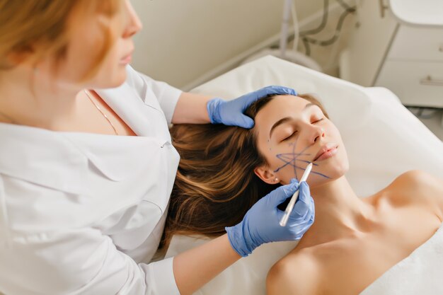 Retrato de mujer hermosa en preparación para el rejuvenecimiento, operación de cosmetología en salón de belleza. Vista desde arriba de las manos del médico en guantes azules dibujando en la cara, botox, belleza