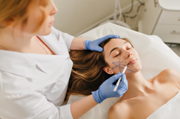 Retrato de mujer hermosa en preparación para el rejuvenecimiento, operación de cosmetología en salón de belleza. Vista desde arriba de las manos del médico en guantes azules dibujando en la cara, botox, belleza