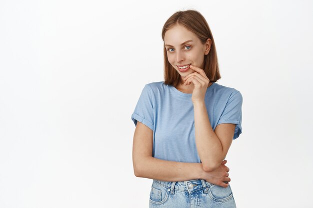 Retrato de mujer hermosa rubia femenina con ojos azules, mirando al frente coqueta, sonriendo coqueta, tiene una idea, pensamiento interesante, de pie contra la pared blanca.