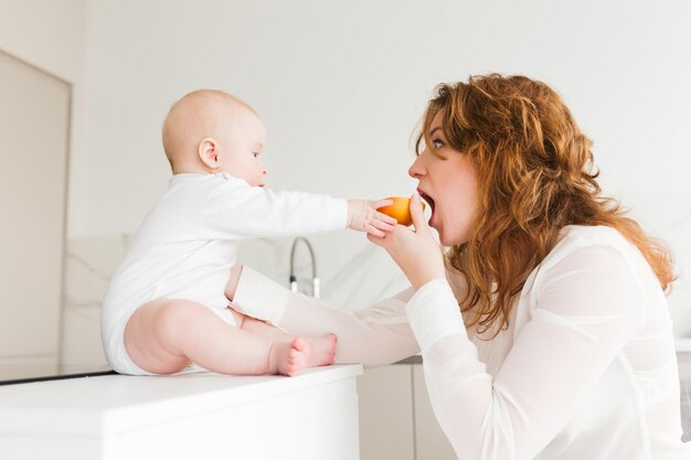 Retrato de una mujer hermosa sentada y comiendo naranja mientras juega alegremente con su lindo bebé