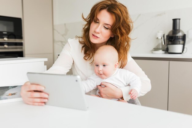 Retrato de una mujer hermosa sentada y sosteniendo a su pequeño bebé en la mano mientras usa su tableta