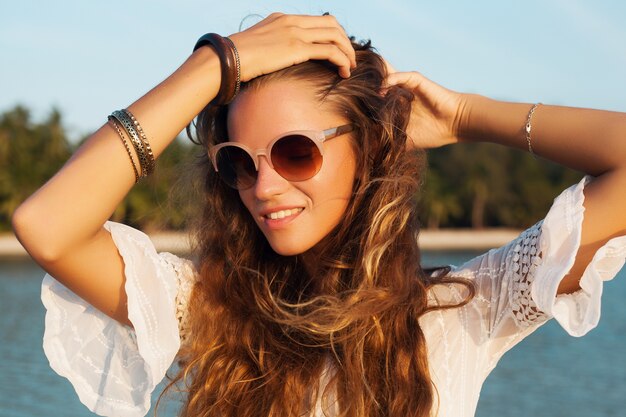 Retrato de mujer hermosa con vestido blanco en playa tropical al atardecer de cerca con elegantes gafas de sol.