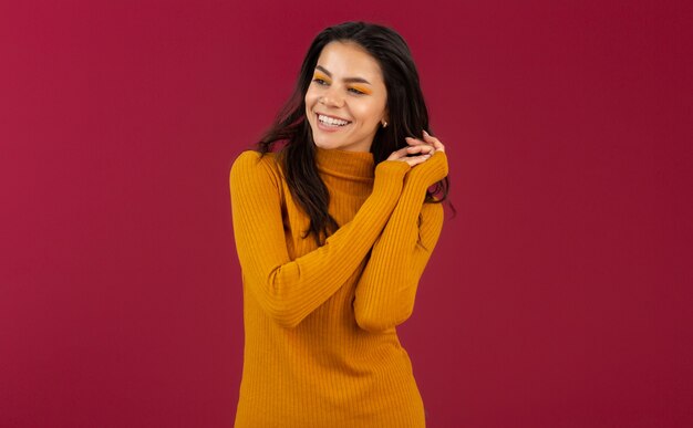 Retrato de mujer hispana morena con estilo muy sonriente en suéter amarillo vestido de moda otoño invierno posando aislado en la pared roja