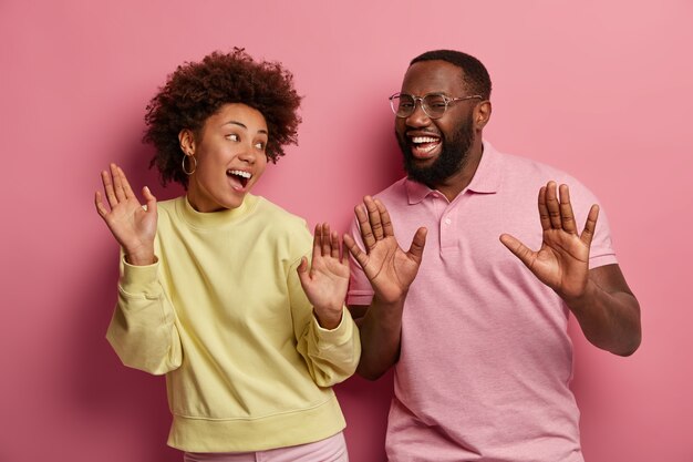 Retrato de mujer y hombre étnicos levantan las palmas de las manos, se sienten optimistas, bailan y se mueven activamente en la fiesta disco, vestidos de manera informal, miran con amplias sonrisas, aislados sobre fondo rosa.