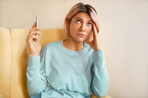 Retrato de mujer independiente joven frustrada con cabello rosado que tiene dolor de cabeza, sosteniendo la mano en su sien mientras habla con un cliente disgustado, moviendo el teléfono a un lado para no escuchar los gritos del otro lado