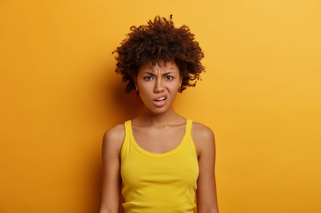 Retrato de mujer insatisfecha de piel oscura sonríe, frunce los labios y se ve con molestia, viste ropa informal, descontento al escuchar algo desagradable, posa en el interior sobre una pared amarilla