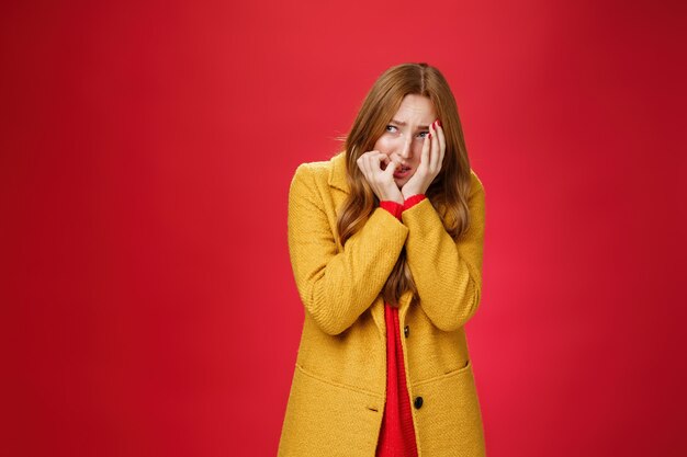 Retrato de mujer insegura conmocionada y asustada con abrigo amarillo que cubre la cara con horror y miedo, mordiéndose las uñas, entrecerrando los ojos y mirando intensamente a la izquierda como víctima asustada de pie en tembloroso, tembloroso.