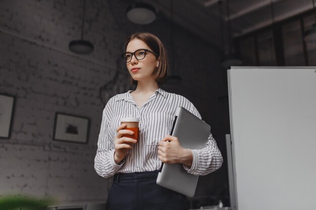 Retrato de mujer inteligente en gafas sosteniendo una taza de té Empleado en traje elegante posando con computadora portátil