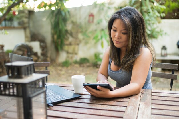 Retrato de mujer japonesa concentrada con tableta en café al aire libre. Hermosa chica comprando o charlando en línea, divirtiéndose, leyendo, trabajando como autónomo. Bebiendo café