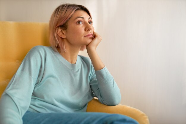 Retrato de mujer joven aburrida con peinado bob rosado sentado en el sofá amarillo en casa con la mano debajo de la mejilla