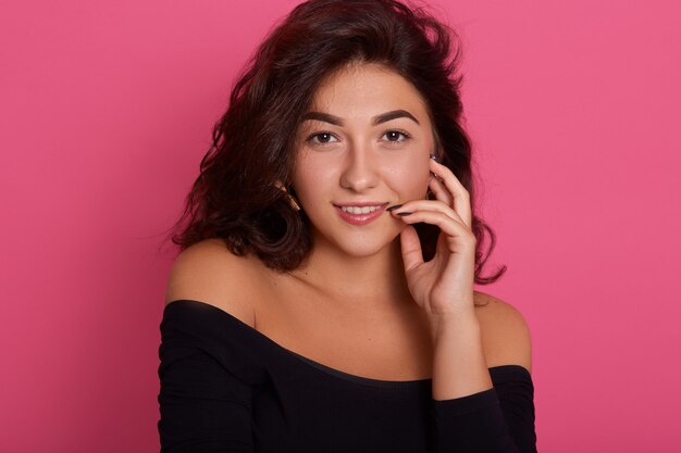 Retrato de mujer joven alegre fresca con sonrisa coqueta, posando aislada sobre pared rosa con el dedo en los labios, mujer de cabello oscuro.