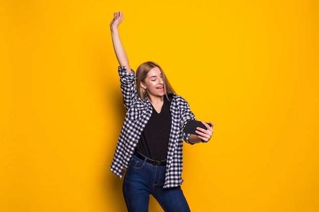 Retrato de una mujer joven alegre que sostiene el teléfono móvil, celebrando que se encuentran aisladas sobre la pared amarilla