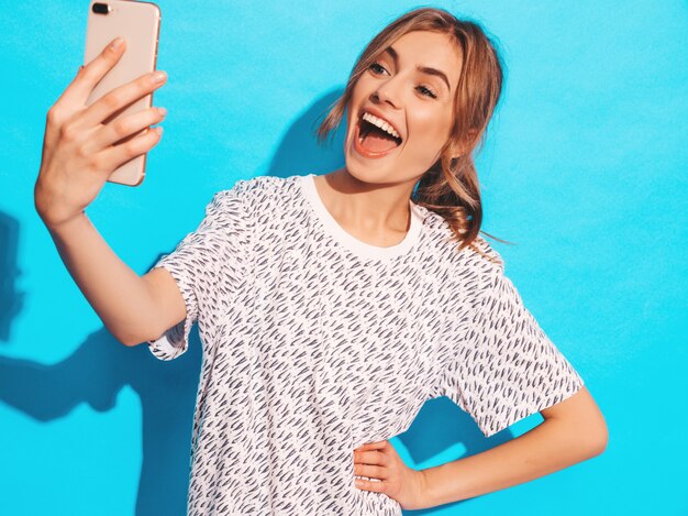 Retrato de mujer joven alegre tomando foto selfie. Hermosa chica con cámara del teléfono inteligente. Modelo sonriente que presenta cerca de la pared azul en estudio. Modelo sorprendido sorprendido