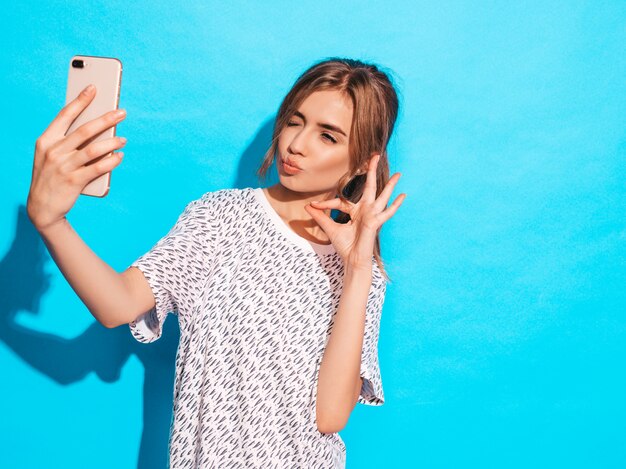 Retrato de mujer joven alegre tomando foto selfie. Hermosa chica con cámara del teléfono inteligente. Modelo sonriente que presenta cerca de la pared azul en estudio. Muestra un signo de ok. Guiños y hace cara de pato