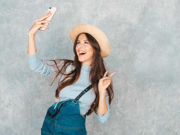 Retrato de mujer joven alegre tomando foto selfie y vistiendo ropa moderna y sombrero. . Muestra signo de paz