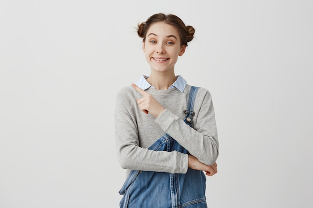 Retrato de mujer joven alegre vistiendo denim señalando con el dedo. Emociones positivas de chica gesticulando en pared blanca proponiendo servicios. Copia espacio