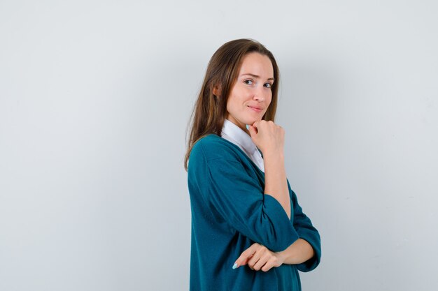 Retrato de mujer joven apoyando la barbilla en la mano en suéter sobre camisa blanca y mirando sensible vista frontal