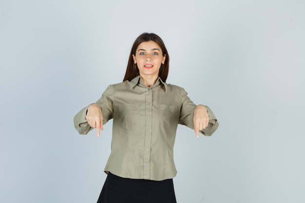 Foto gratuita retrato de mujer joven apuntando hacia abajo en camisa, falda y mirando alegre vista frontal