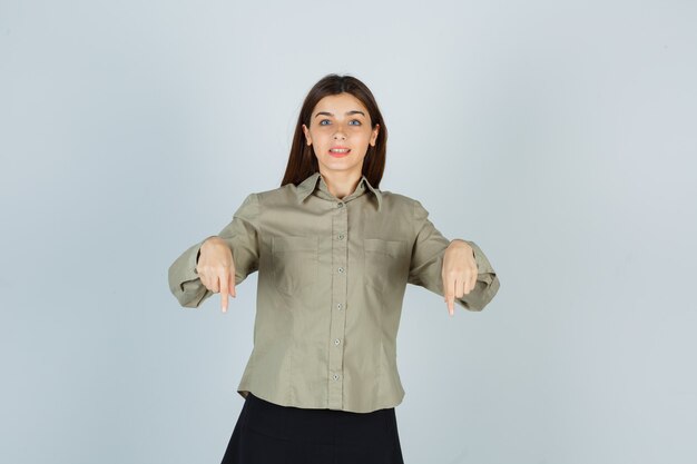 Retrato de mujer joven apuntando hacia abajo en camisa, falda y mirando alegre vista frontal