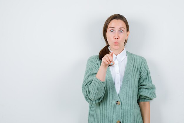 Retrato de mujer joven apuntando hacia adelante en blusa, chaqueta de punto y mirando sorprendido vista frontal