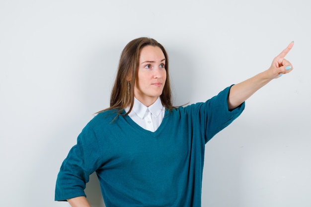 Retrato de mujer joven apuntando hacia afuera en suéter sobre camisa y mirando enfocado vista frontal