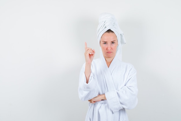 Retrato de mujer joven apuntando hacia arriba en bata de baño blanca, toalla y mirando vacilante vista frontal