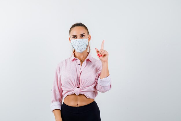 Retrato de mujer joven apuntando hacia arriba en camisa, pantalones, máscara médica y mirando confiada vista frontal