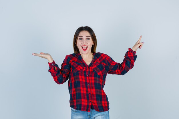 Retrato de mujer joven apuntando hacia arriba, extendiendo la palma hacia un lado en camisa a cuadros y mirando sorprendido vista frontal