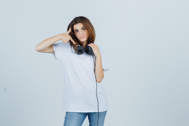 Retrato de mujer joven apuntando a los auriculares en camiseta blanca, jeans y mirando atractiva vista frontal