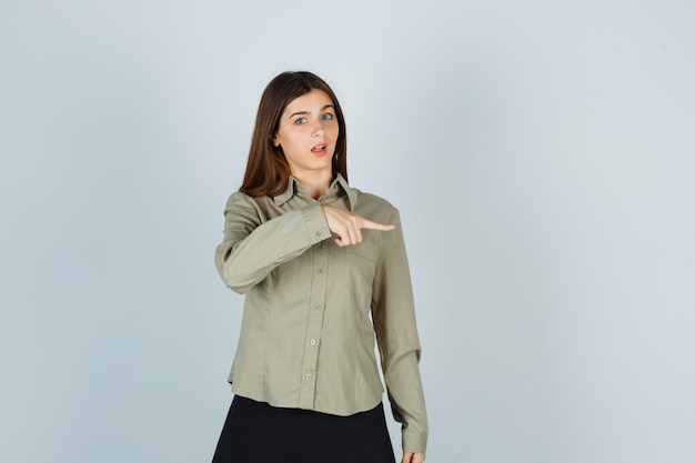 Foto gratuita retrato de mujer joven apuntando a la derecha en camisa, falda y mirando sorprendido vista frontal