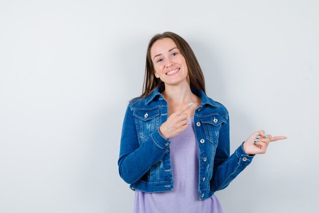 Retrato de mujer joven apuntando a la derecha en camiseta, chaqueta y mirando alegre vista frontal