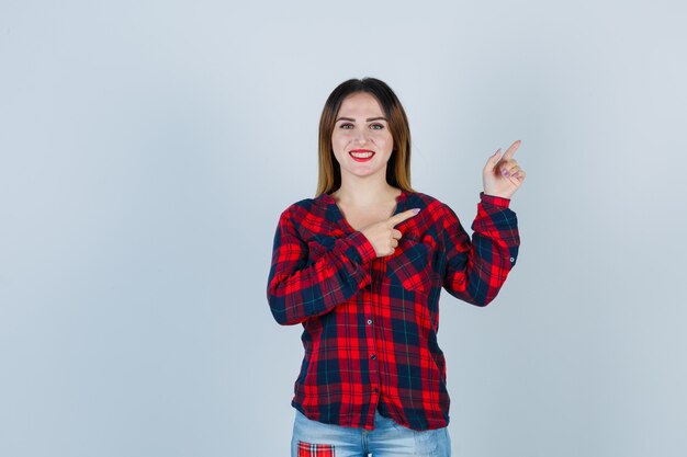 Retrato de mujer joven apuntando a la esquina superior derecha en camisa a cuadros y mirando alegre vista frontal