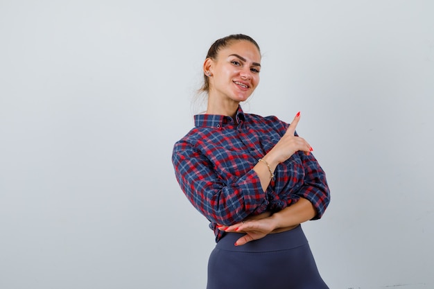 Foto gratuita retrato de mujer joven apuntando a la esquina superior derecha en camisa a cuadros, pantalones y mirando feliz vista frontal