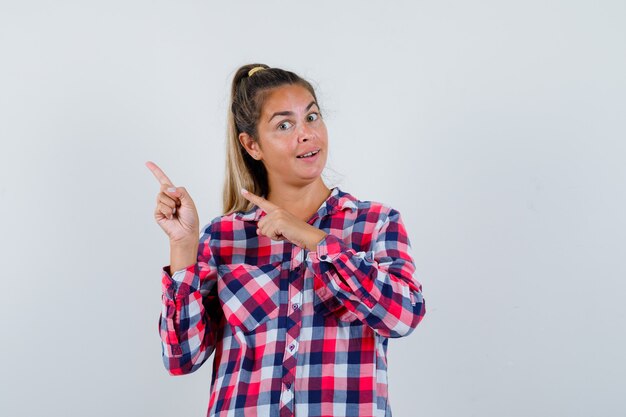 Retrato de mujer joven apuntando a la esquina superior izquierda en camisa casual y mirando feliz vista frontal