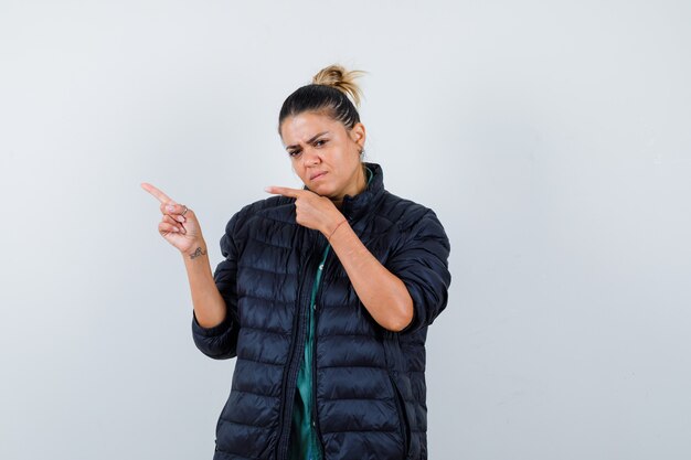 Retrato de mujer joven apuntando a la esquina superior izquierda en chaqueta acolchada y mirando indeciso vista frontal