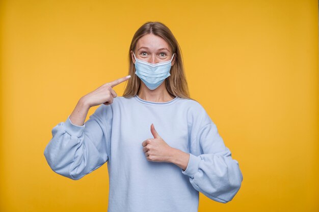 Retrato de una mujer joven apuntando a su máscara médica y mostrando los pulgares hacia arriba